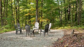 A forest setting with a gravel area and four Adirondack chairs arranged around a central table.