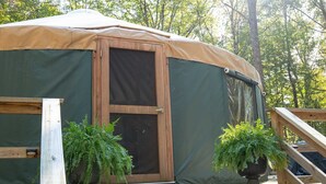 Eine große, runde, überdachte Außenkonstruktion mit einer Tür und Fenstern, umgeben von einer Holzterrasse und zwei Kübelpflanzen.