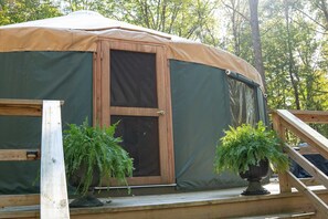 Una estructura circular grande y cubierta al aire libre, con puerta y ventanas, rodeada por una terraza de madera y dos plantas en macetas.