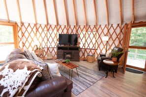 A cozy living room with a sofa, a TV, a fireplace, and a view of the outdoors. - Arbor Ridge Yurt- Hocking Hills Yurts and Cabins (Laurelville)
