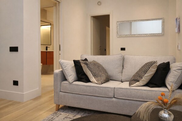 A living room with a grey sofa, black and white patterned pillows, a mirror on the wall, and a vase with dried plants on a side table.