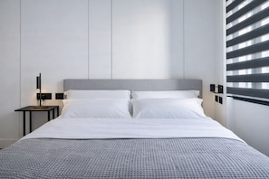Quarto de hotel moderno com uma cama grande, uma mesinha de cabeceira com abajur e uma janela com persianas.