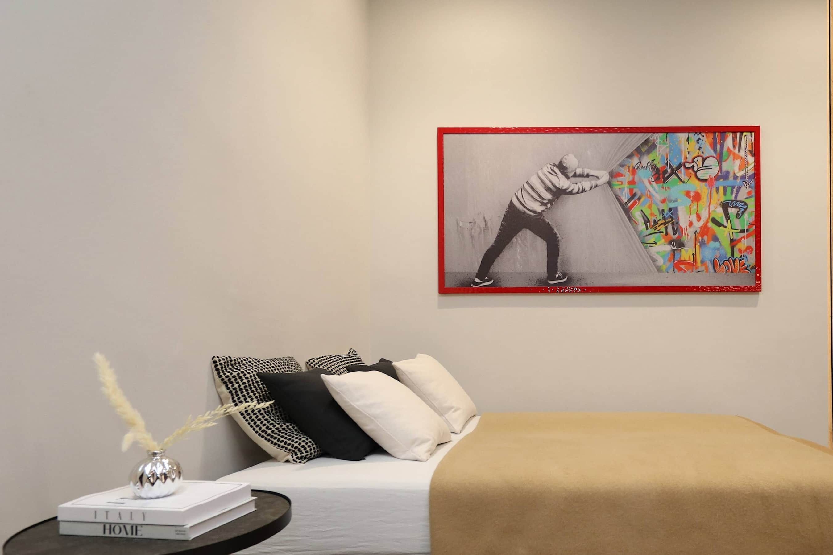 Quarto com cama, almofadas, mesa de cabeceira com livros, um vaso e uma obra de arte emoldurada na parede.