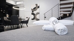 A modern hotel room with a neatly made bed, a dining area with chairs, and a staircase.