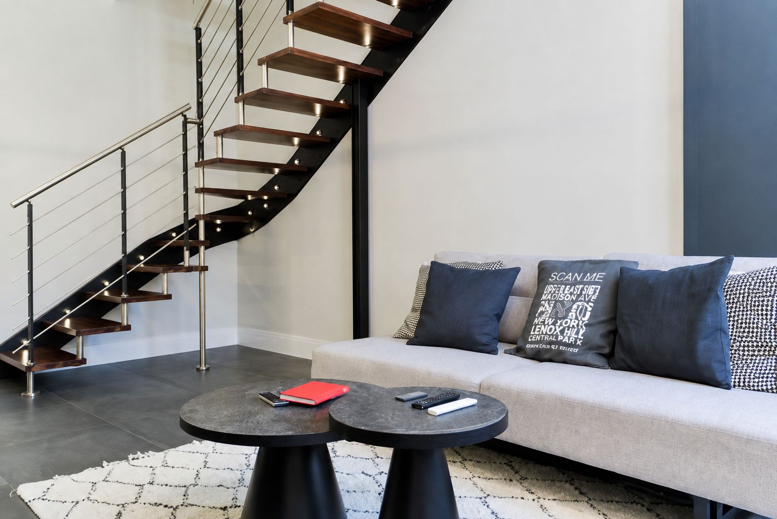 A modern living room with a curved staircase, a sofa with patterned cushions, and a round coffee table with books and remotes.