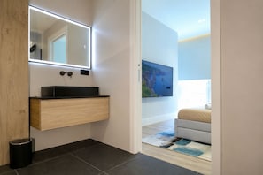 Un baño moderno con tocador de madera, un espejo con iluminación integrada, un lavabo negro y un cubo de basura negro.