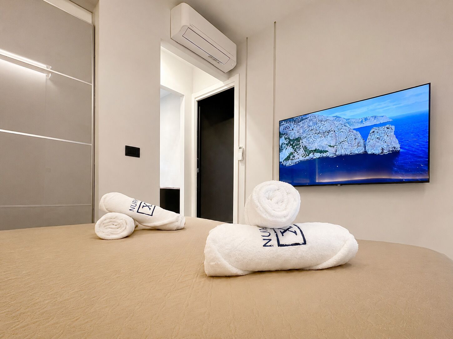 A hotel room with a bed, a wall-mounted TV displaying a mountainous landscape, and two towels with a logo on them.
