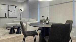 Uma sala de jantar moderna com uma mesa redonda, cadeiras pretas e uma luminária pendente. Há duas pinturas abstratas emolduradas na parede, um abajur e um sofá ao fundo.