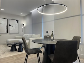 Un comedor moderno con una mesa redonda, sillas negras y una lámpara colgante. En la pared, hay dos cuadros abstractos enmarcados, una lámpara de pie y un sofá al fondo.