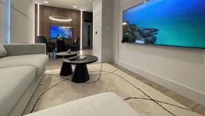 Uma sala de estar moderna com uma grande televisão de tela plana, um sofá, uma mesa de centro e uma área de jantar com cadeiras e uma televisão.
