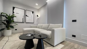 Uma sala de estar moderna com um sofá branco, duas mesas de centro pretas, um vaso com planta e arte abstrata na parede.