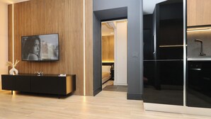 A modern living room with a large flat-screen TV mounted on a wooden panel wall, a black console table with decorative items, and a view into the adjacent bedroom.