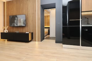Una sala moderna con un televisor de pantalla plana grande montado en una pared de paneles de madera, una consola negra con objetos decorativos, y vista al dormitorio contiguo.