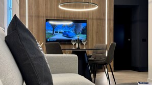 A modern living room with a sofa, a dining table with chairs, a TV mounted on the wall, and a wine glass and bottle on the table.