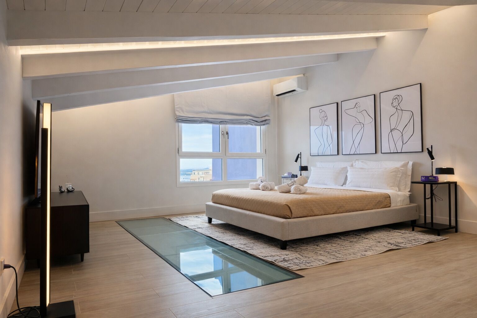 A modern bedroom with a large bed, a glass floor-to-ceiling window, and artwork on the walls.