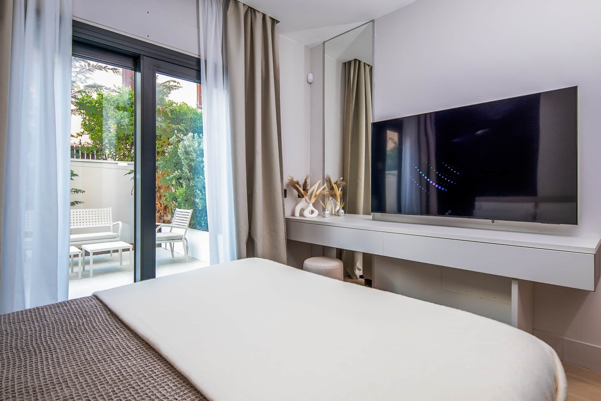 Ein modernes Hotelzimmer mit einem großen Bett, einem flachbildfernseher an der Wand, einer Glastür, die zu einem Sitzbereich im Freien führt, und einer minimalistischen Einrichtung.