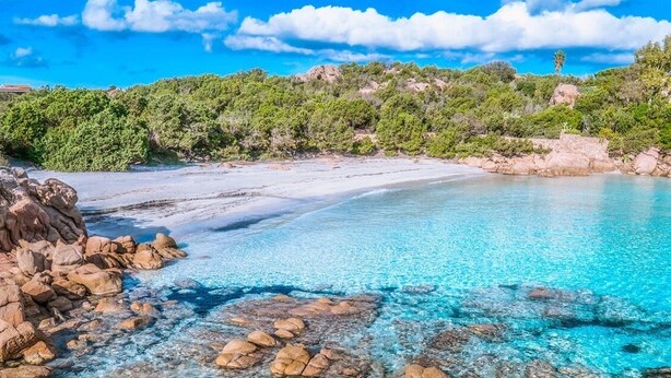 A coastal beach with clear turquoise waters, rocky shoreline, and dense greenery in the background.