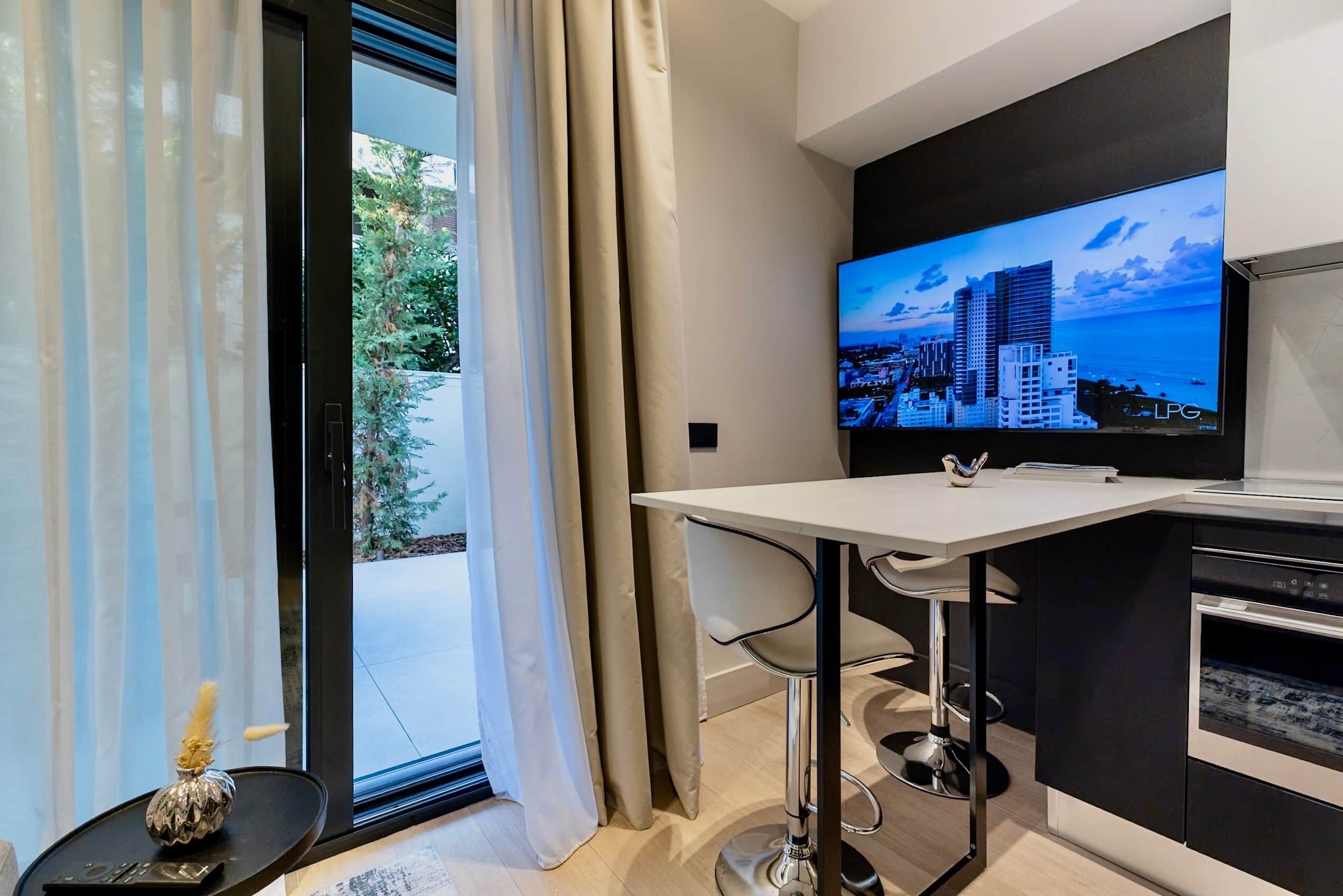 Una cocina moderna con una televisión grande mostrando un paisaje urbano, un área de comedor con una mesa blanca y sillas negras, y vistas a zonas verdes por la ventana.