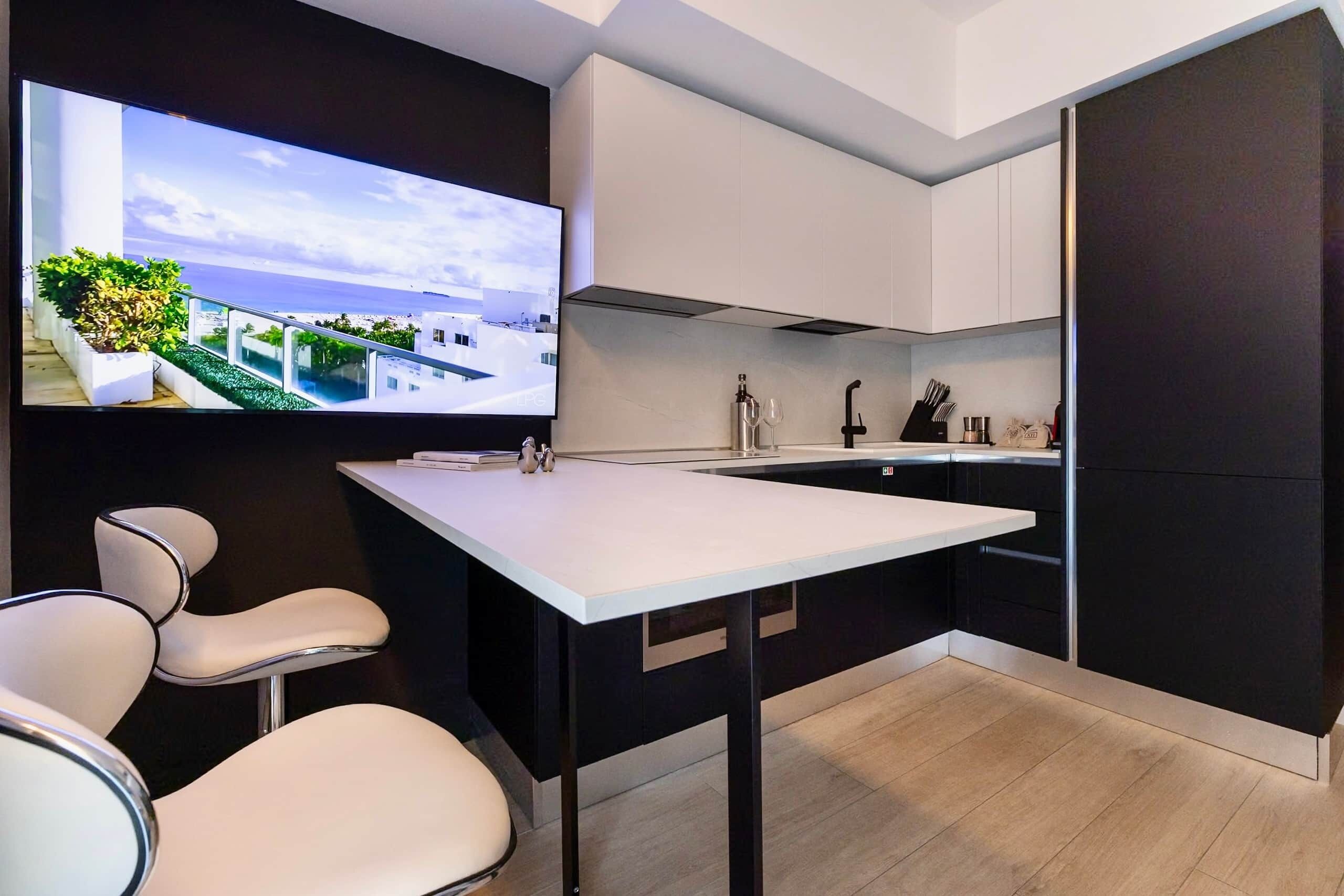 A modern kitchen with a large island, white chairs, and a TV displaying a scenic view.