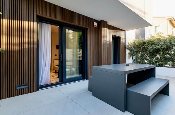 Un espacio exterior moderno con una banca integrada y una mesa, junto a una puerta corrediza de vidrio que da a una habitación.