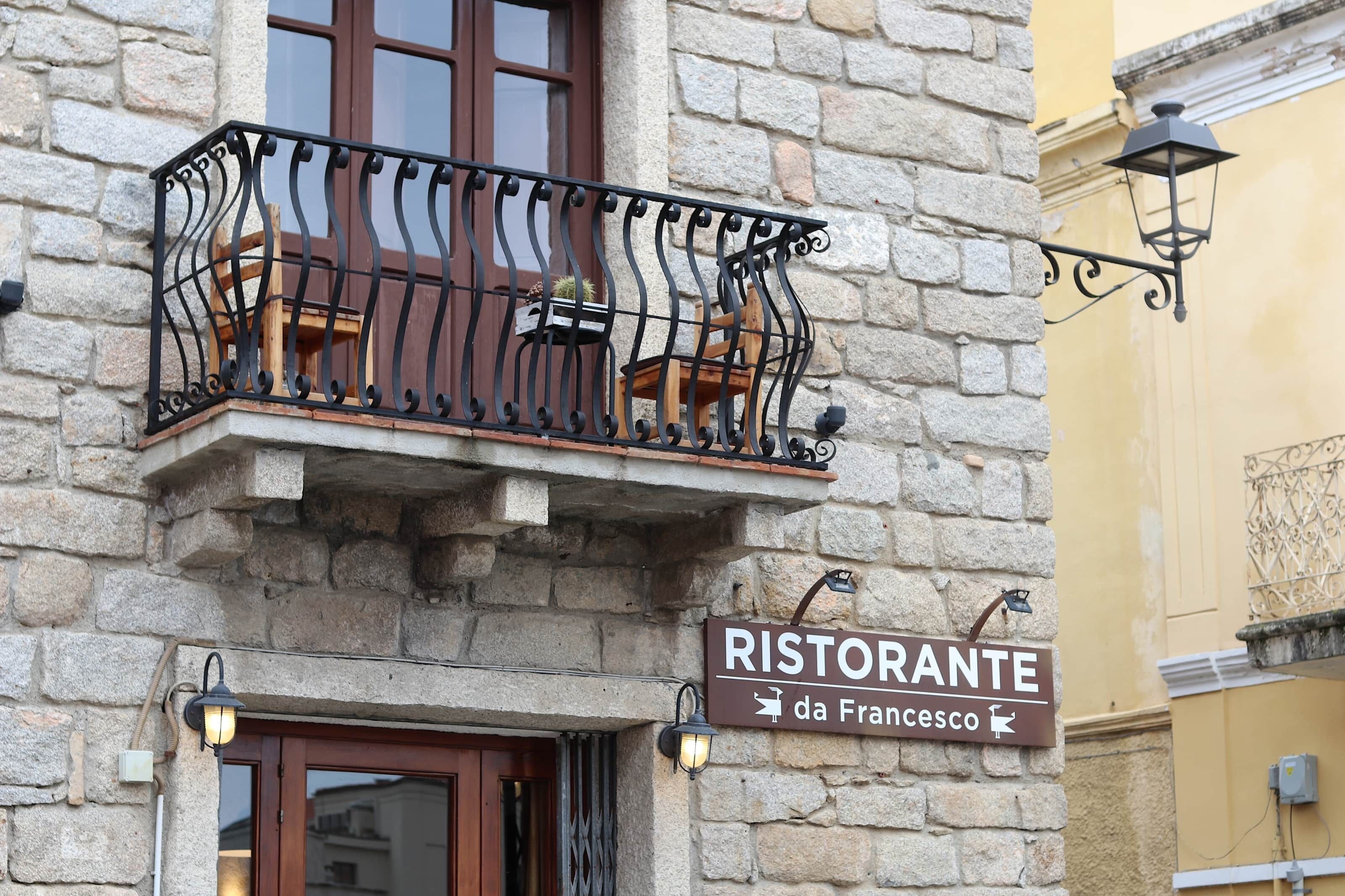 Un bâtiment en pierre doté d’un balcon orné de balustrades en fer forgé et de chaises en bois. On peut y voir une enseigne indiquant « RISTORANTE da Francesco ».