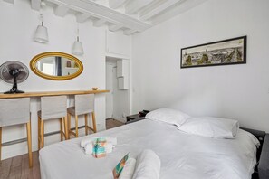 A hotel room with a bed, a desk with chairs, a fan, a mirror, and a framed picture on the wall.