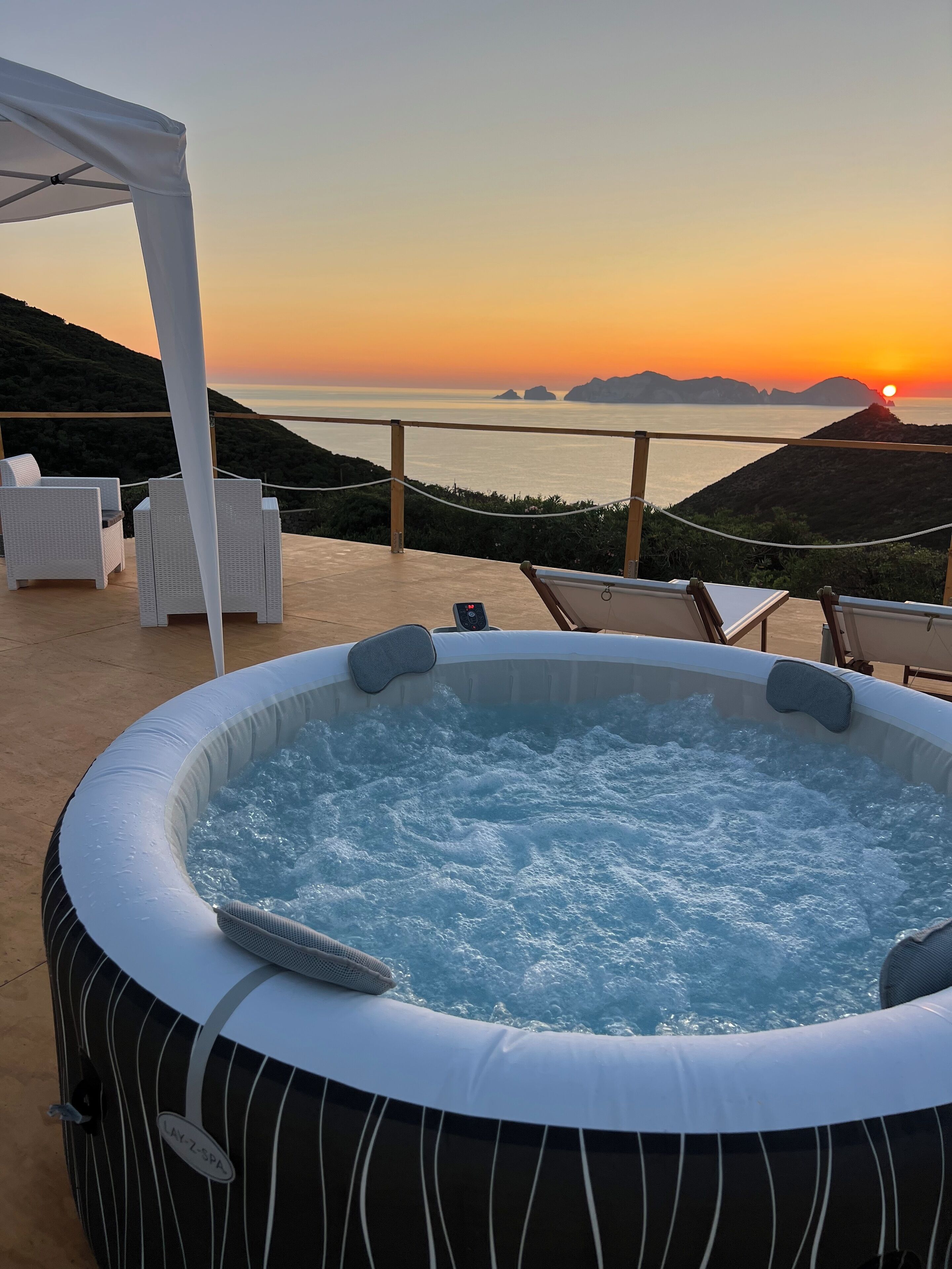 A hot tub with a view of the ocean at sunset, surrounded by lounge chairs and a pergola.