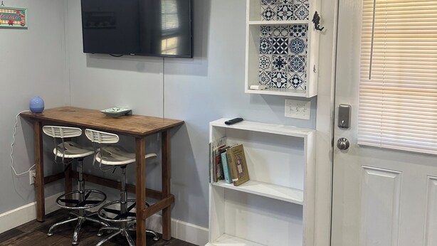 Quarto com mesa de madeira, dois taburetes brancos, televisão de tela plana fixada na parede, porta branca com tapete e uma prateleira com azulejos estampados.