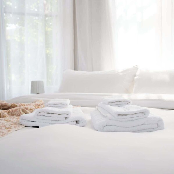 A neatly made bed with white linens and towels, a bedside lamp, and sheer curtains allowing natural light.