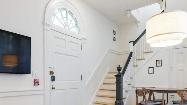 A living room with a staircase, a TV mounted on the wall, and a dining area with chairs and a table.