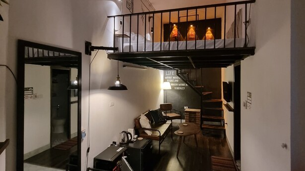 Un espacio habitable compacto con un entrepiso que alberga un dormitorio sobre un área de comedor. El entrepiso cuenta con una cama y una pequeña zona de estar con una mesa y sillas. El área de comedor tiene una mesa con sillas y una cafetera sobre la mesa.