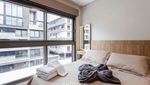A modern hotel room with a large window, a bed with a beige quilt, a wooden headboard, and a view of other buildings. - Beautiful apt Less Than 5min From the Beach Vrn302 (Florianópolis)