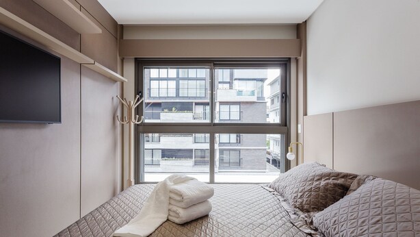 A bedroom with a bed, a TV mounted on the wall, a window with a view of buildings, and a coat rack. - Beautiful apt Less Than 5min From the Beach Vrn302 (Florianópolis)