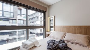 A modern hotel room with a large window, a bed with a beige quilt, a wooden headboard, and a view of other buildings.