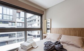 A modern hotel room with a large window, a bed with a beige quilt, a wooden headboard, and a view of other buildings.