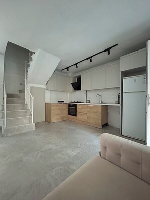 A modern kitchen with a central island, white cabinets, and a built-in oven. A staircase leads to a landing with a beige sofa. - New Villa Steps from Long Beach, Kusadasi (Aydın)