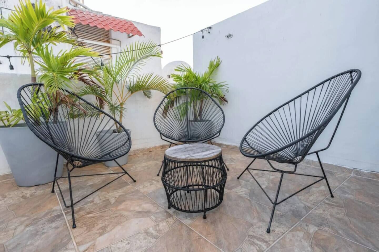 Une terrasse avec trois chaises à armature métallique noire et une table ronde, entourée de plantes en pot et d’un mur blanc.
