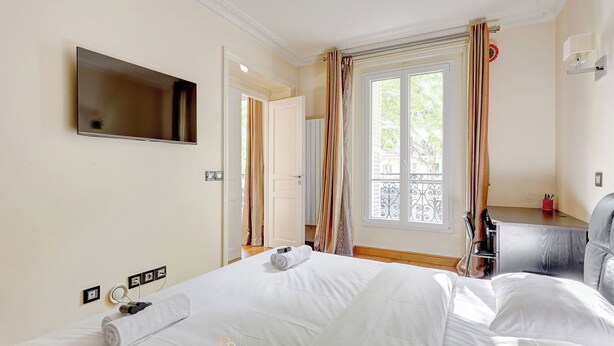 A hotel room with a bed, a TV mounted on the wall, a window with curtains, and a desk with a chair.