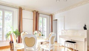 Ein Zimmer mit Klavier, einem Esstisch mit Stühlen und großen Fenstern mit Vorhängen.