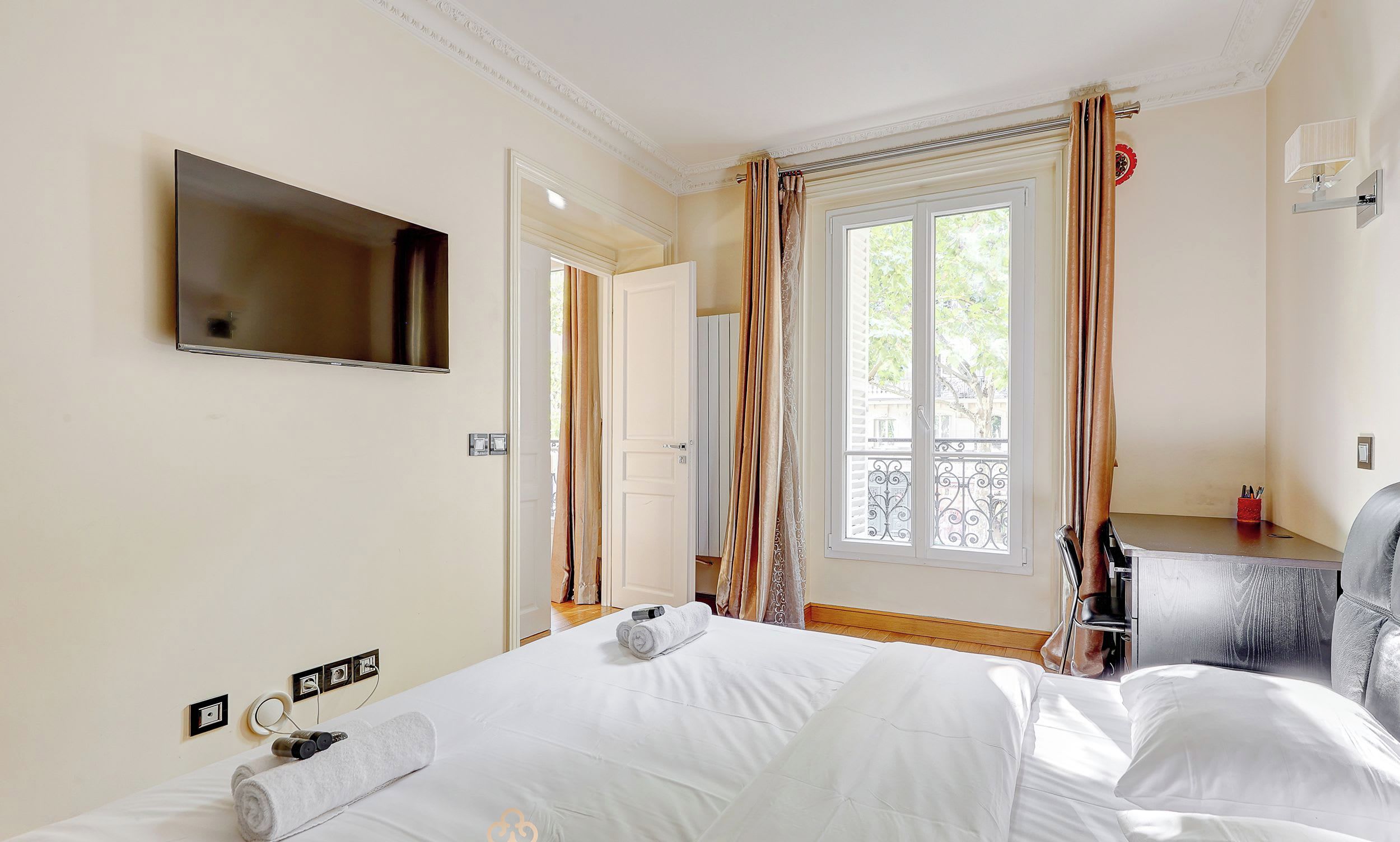 A hotel room with a bed, a TV mounted on the wall, a window with curtains, and a desk with a chair.