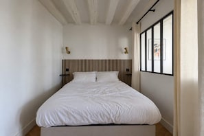 A bedroom with a bed, a headboard with vertical lines, two wall-mounted light fixtures, and a window with curtains. - Cocon parisien • Rue de la Verrerie (Paris)