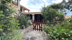 Un sendero de piedra conduce a una verja de madera, rodeado de abundante vegetación y un edificio con techo de tejas.