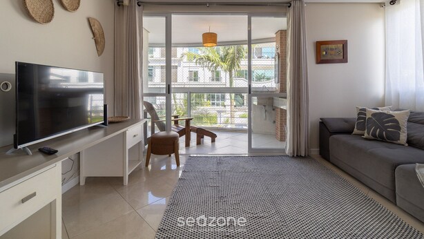 A modern living room with a flat-screen TV, a sofa, a dining area with a table and chairs, and a balcony with a view of buildings and trees. - Beautiful apt 3 Minutes From Jurere Beach Vma101 (Florianópolis)