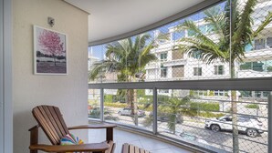 A balcony with a wooden chair and a colorful cushion, overlooking a street with palm trees and parked cars. - Beautiful apt 3 Minutes From Jurere Beach Vma101 (Florianópolis)
