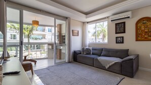 A modern living room with a grey sofa, a coffee table, and a large window offering a view of a building and palm trees. - Beautiful apt 3 Minutes From Jurere Beach Vma101 (Florianópolis)