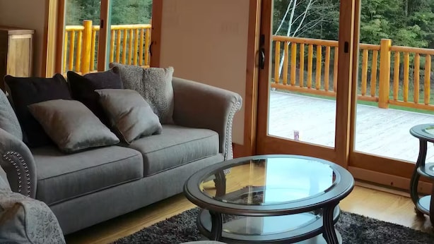 A spacious living room with a high ceiling and large windows, a grey sofa, a glass coffee table, and a wooden deck visible through the glass doors. - Modern 3BR/4BA Windham chalet w/ hot tub, views & mins to ski, bars, dining! (Windham)