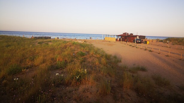 A beach with a building, a fenced area, and a dirt path leading to the water.