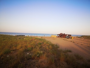 A beach with a building, a fenced area, and a dirt path leading to the water.
