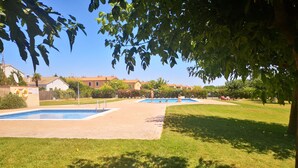 A swimming pool area with two pools, surrounded by grass and trees, with buildings in the background.