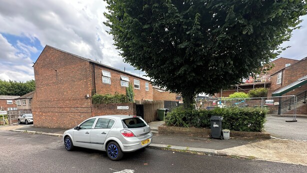 Una calle residencial con un coche blanco estacionado en la banquina, un edificio de ladrillo y un árbol.